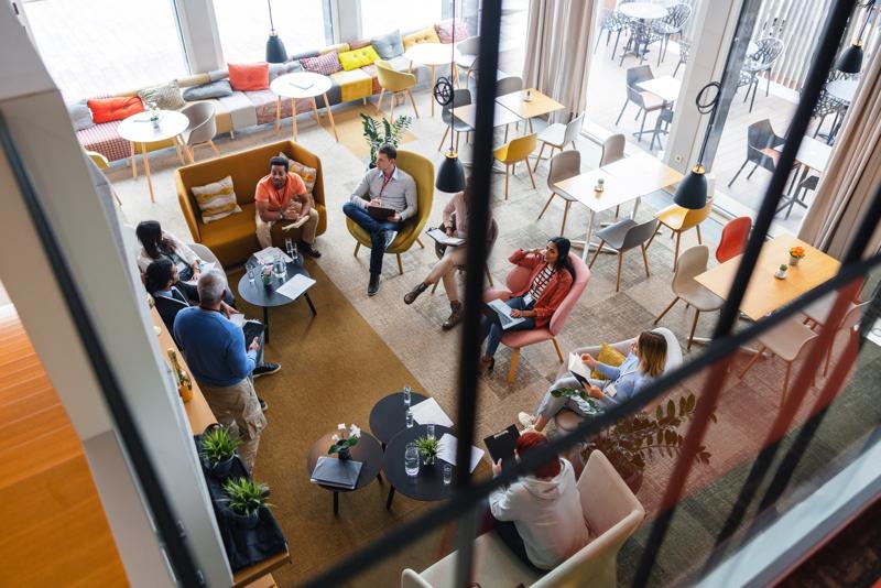 A diverse group of corporate employees sitting in a circle and brainstorming the options for strategy improvement. The meeting is led by the black mature adult CEO. The atmosphere is relaxed since they are located in a colourful and cosy office cafeteria.