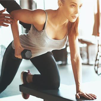 Personal Trainer Closeup shot of two women doing dumbbell rows at a local gym. Leaning over workout bench and looking at a large mirror that is not in the frame.They are assisted by two unrecognizable male coaches, one of them not visible at all.