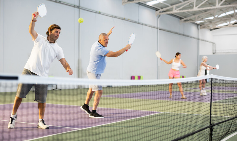 Portrait of sporty adult man playing doubles pickleball with experienced aged partner on indoor court, ready to hit ball. Sport and active lifestyle concept