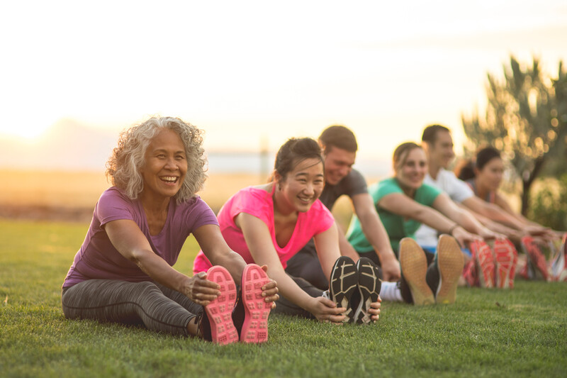 A group of adults attending a fitness class outdoors are doing leg stretches. The participants are arranged in a line. The focus is on a mature ethnic woman who is smiling toward the camera.