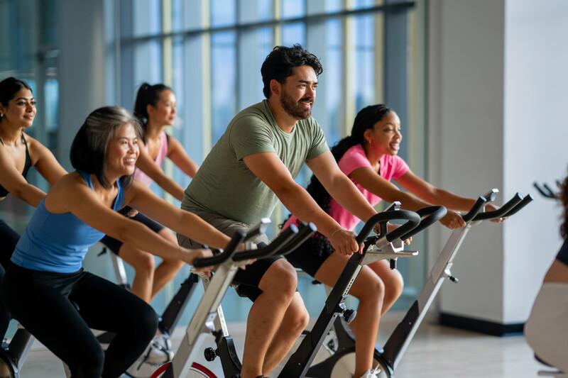 A small group of mature adults are seen sitting on stationary bikes, as they participate in a cycling fitness class together.  They are each dressed comfortably and are focused on the workout.