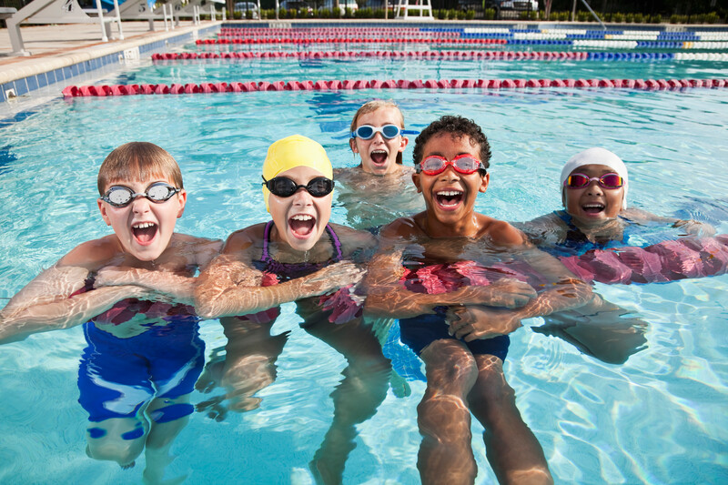 A multiracial group of five children having fun in a swimming pool, hanging onto a lane rope, looking at the camera, laughing and shouting.  The three boys and two girls are 9 to 12 years old.  They are wearing swimsuits and goggles, and the girls have on swim caps.